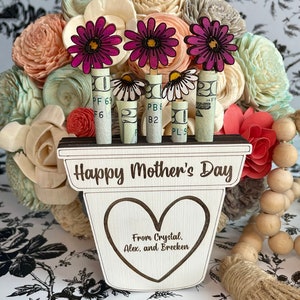 May include: A wooden planter with the words "Happy Mother's Day" and "From Crystal. Alex, and Brecken" engraved on it. Three pink flowers are attached to dollar bills that are inserted into the planter.
