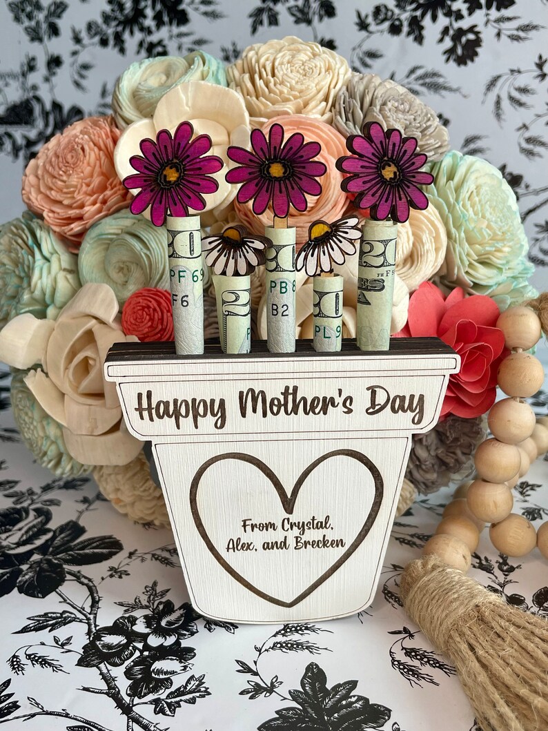 May include: A wooden planter with the words "Happy Mother's Day" and "From Crystal. Alex, and Brecken" engraved on it. Three pink flowers are attached to dollar bills that are inserted into the planter.