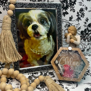 May include: A wooden box with a hexagonal shape and a clear lid. The box has a brown wood finish and is engraved with the name "Chloe" and the dates "2007-2021". A red heart is visible inside the box. A small white tassel hangs from the lid. The box is sitting on a white and black floral background.
