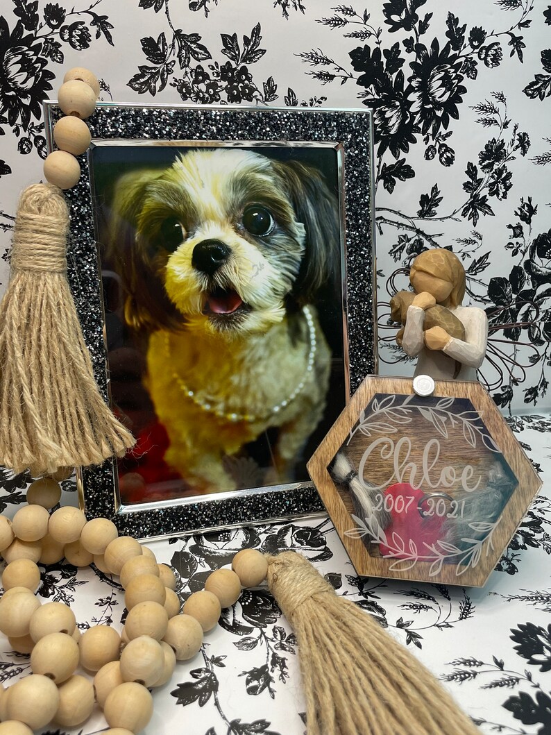 May include: A wooden box with a hexagonal shape and a clear lid. The box has a brown wood finish and is engraved with the name "Chloe" and the dates "2007-2021". A red heart is visible inside the box. A small white tassel hangs from the lid. The box is sitting on a white and black floral background.