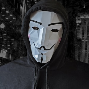 Puede incluir: Una persona con una sudadera con capucha negra con la capucha puesta sobre la cabeza. Lleva una máscara blanca con un diseño geométrico y un bigote negro.