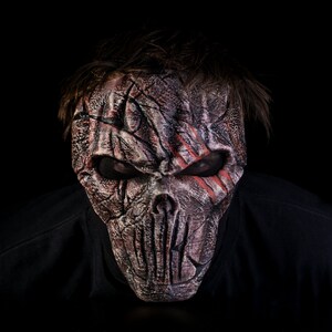 May include: A person wearing a scary, detailed, gray and red skull mask with a black background.