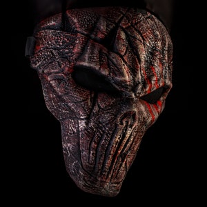 May include: A black and red Halloween mask with a textured, cracked, and weathered design. The mask has a skull-like shape with a single eye hole and red streaks of paint.