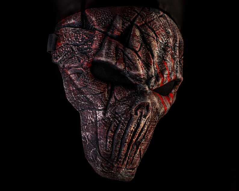 May include: A black and red Halloween mask with a textured, cracked, and weathered design. The mask has a skull-like shape with a single eye hole and red streaks of paint.