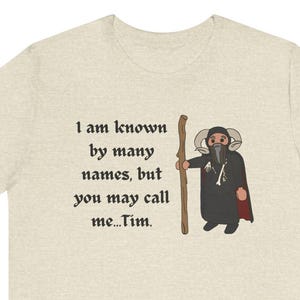 May include: Beige t-shirt with the text "I am known by many names, but you may call me...Tim." A cartoon illustration of a wizard holding a staff is to the right of the text. The wizard wears a black robe and a red cape.