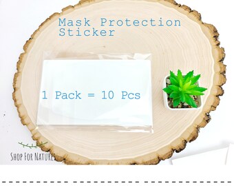 May include: A close-up of a package of 10 mask protection stickers. The stickers are white and are packaged in a clear plastic bag. The text "1 Pack = 10 Pcs" is printed on the package. The package is sitting on a wooden surface.
