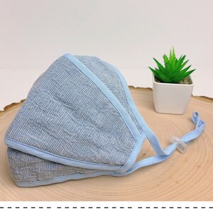 May include: A light blue face mask with a textured pattern and a white adjustable strap. The mask is lying on a wooden surface.