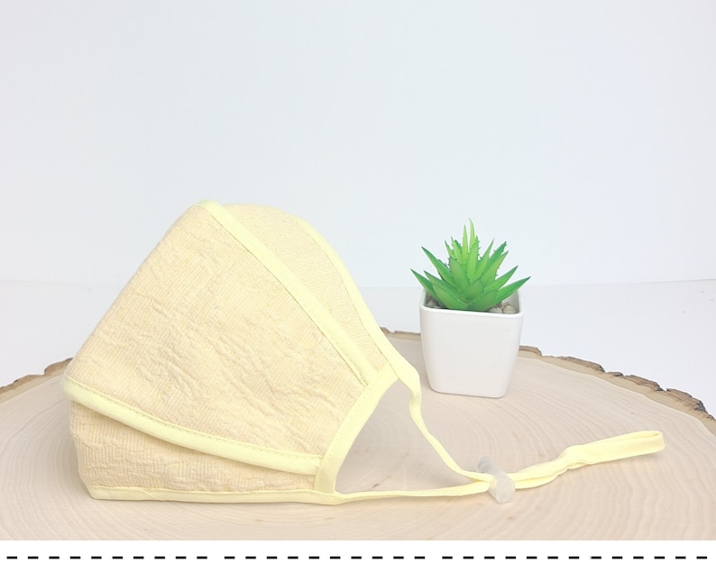May include: A pale yellow face mask with a light yellow trim and adjustable straps. The mask is made of a soft, textured fabric.