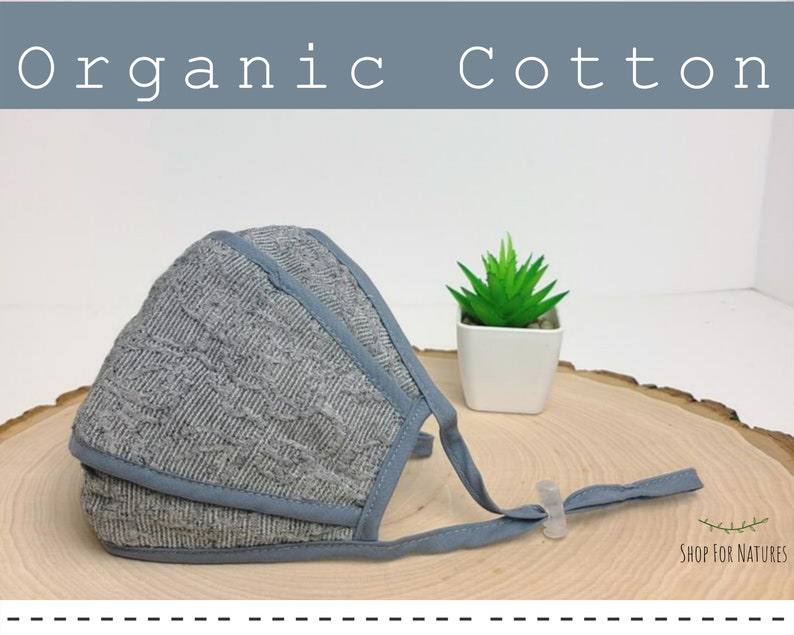 May include: A gray organic cotton face mask with adjustable ear loops and a nose wire. The mask is on a wooden surface with a small potted plant in the background. The text "Shop for Natures" is visible in the bottom right corner.
