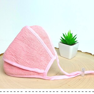 May include: Pink face mask with a textured pattern and adjustable straps. The mask is folded and lying on a wooden surface.