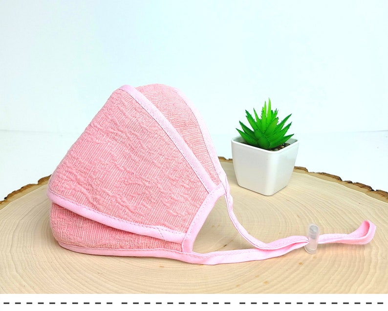 May include: Pink face mask with a textured pattern and adjustable straps. The mask is folded and lying on a wooden surface.
