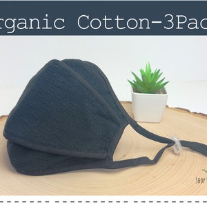 May include: A black organic cotton face mask with adjustable ear loops and a nose wire. The mask is folded in half and has a textured surface. The mask is on a wooden surface with a small plant in a white pot behind it. The text "Shop for Natures" is visible in the bottom right corner of the image.