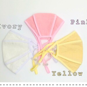 May include: Three fabric face masks in ivory, pink, and yellow. The masks are made of a soft, textured material and have a simple, rectangular shape.