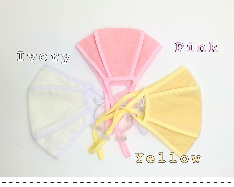 May include: Three fabric face masks in ivory, pink, and yellow. The masks are made of a soft, textured material and have a simple, rectangular shape.