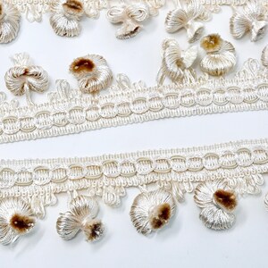 May include: Ivory trim with a delicate woven design and small, fluffy tassels. The tassels have a brown center.