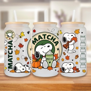 Op de afbeelding: Drie glazen bekers van matglas met bamboe deksels en rietjes, voorzien van Snoopy- en Woodstock-afbeeldingen, herfstbladeren, pompoenen en eikels. Het ontwerp bevat de tekst "MATCHA" in een cirkelvormig logo en een checklist voor diverse opties.