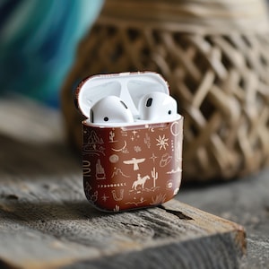 Op de afbeelding: Een bruine AirPod-hoes met een wit en gouden ontwerp met verschillende westerse symbolen, waaronder cactussen, hoefijzers en een cowboyhoed.