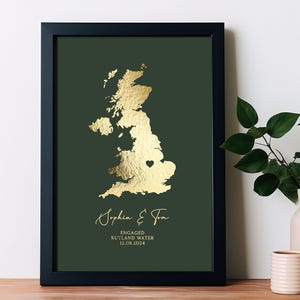 Puede incluir: Impresión artística enmarcada con un mapa dorado del Reino Unido sobre un fondo verde oscuro. La impresión incluye un corazón que marca una ubicación, los nombres "Sophia & Tom" y el texto "Engaged Rutland Water 12.08.2024".