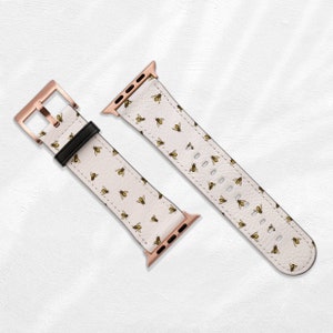 Könnte beinhalten: Ein weißes Lederarmband für die Apple Watch mit einer goldenen Schließe und einem Muster aus kleinen Bienen. Das Armband ist abgebildet, ohne an einer Uhr befestigt zu sein.