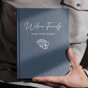 Puede incluir: Un cuaderno de puntuación de juegos de color azul marino con el texto "Wilson Family" en escritura elegante. El libro también presenta el texto "GAME NIGHT SCORES" y un gráfico de naipes. El libro está en manos de alguien.