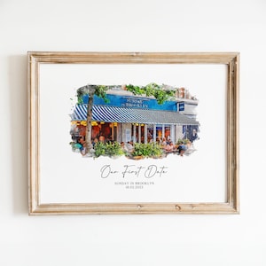 May include: A framed watercolour print depicting a restaurant exterior. The building has a blue facade and a striped awning. The text "Sunday in Brooklyn" is displayed. The print also includes the text "Our First Date" and the date 18.02.2023.