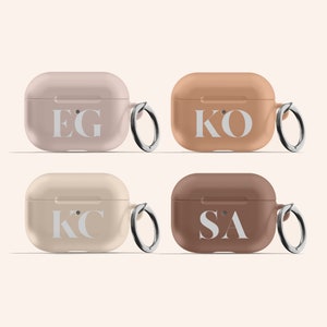 May include: Four Airpod cases in different colors with personalized initials. The cases are beige, light brown, tan, and brown. Each case has a silver ring attached to it.