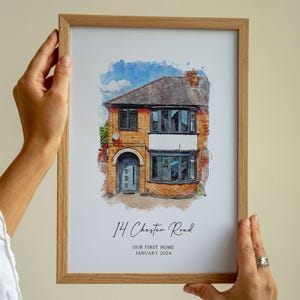 Op de afbeelding: Een ingelijste aquarelprint van een bakstenen huis met twee verdiepingen, met een blauwe deur en ramen. De print bevat het adres "14 Chester Road" en de tekst "OUR FIRST HOME JANUARY 2024". De lijst is van lichtgekleurd hout.