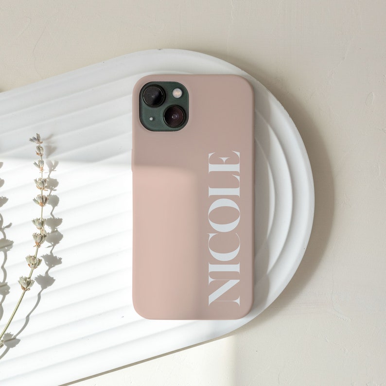 Puede incluir: Funda de tel&eacute;fono rosa p&aacute;lido con el nombre "NICOLE" impreso en blanco verticalmente. La funda est&aacute; sobre una bandeja blanca ovalada. Se ven ramitas de lavanda seca a la izquierda.