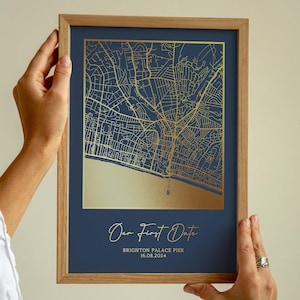 Op de afbeelding: Een ingelijste kaartafdruk met een goudfolie ontwerp op een marineblauwe achtergrond. De kaart toont een gedetailleerde stratenkaart, met de tekst "Our First Date" en locatiegegevens. De lijst is gemaakt van lichtgekleurd hout.