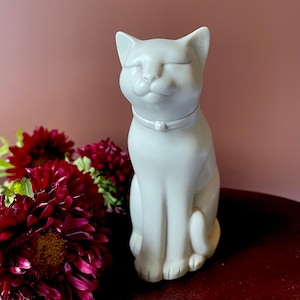 May include: A white ceramic cat figurine with closed eyes and a small collar. The cat is sitting upright, with its paws neatly tucked. Deep red flowers are in the foreground, and the background is a solid pink.