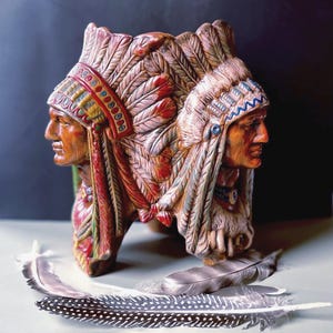 May include: A detailed sculpture of two Native American chiefs facing opposite directions, each wearing a feathered headdress. The sculpture is painted in shades of brown, red, and blue. Several feathers are placed in front of the sculpture.