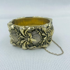 May include: A gold-toned bangle bracelet with an intricate floral design. The bracelet has a chain attached to the clasp.