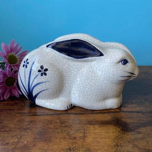 May include: A ceramic rabbit figurine with a crackle glaze finish. The rabbit is white with blue accents on the ears and floral designs on the side. The figurine is sitting on a wooden surface with pink flowers in the background.