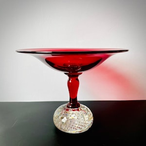 May include: A vibrant red glass pedestal bowl with a broad, shallow top. The stem is solid red, and the base is a clear glass orb with tiny bubbles. The bowl is likely used for serving fruit or displaying decorative items.