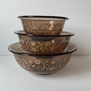 May include: A set of three brown glass mixing bowls stacked on top of each other. Each bowl has a black rim and is decorated with a floral and heart pattern in a lighter brown color. The bowls are nested together.