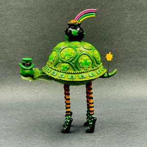 May include: A whimsical St. Patrick's Day figurine. The green turtle-like figure has a shamrock-patterned shell, striped legs, and black shoes. It holds a leprechaun hat and a star wand. A pot of gold with a rainbow sits atop its head.
