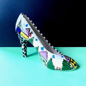 May include: A white high-heeled shoe with a whimsical painted design. The shoe features a black and white checkered heel and is decorated with colorful houses, trees, and floral accents. The interior of the shoe is white with a black dotted trim.