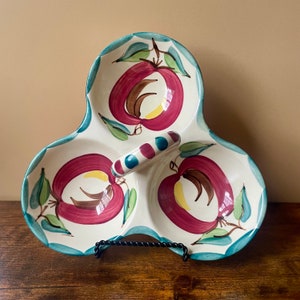 May include: A white ceramic serving dish with three sections, each featuring a hand-painted red apple with green leaves. The dish has a blue and red striped handle.