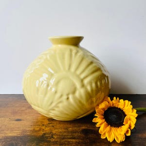 May include: A pale yellow ceramic vase with a textured, sunburst-like design. The vase is round with a small opening at the top. A bright yellow sunflower with a dark center rests beside the vase on a wooden surface.
