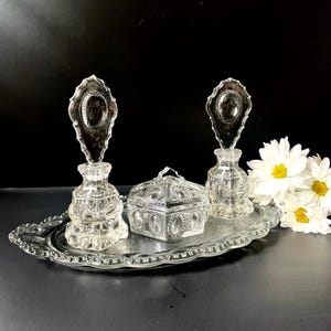 May include: A clear glass vanity set consisting of an oval tray with a scalloped edge, two perfume bottles with tall decorative stoppers, and a lidded hexagonal trinket box. Each piece features a repeating embossed oval pattern in the glass.