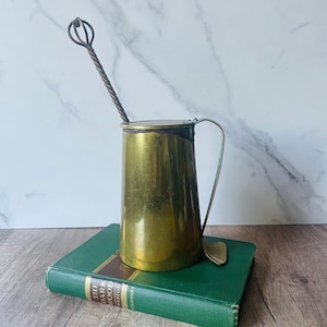 May include: A brass pitcher with a handle and lid, and a long, twisted metal spoon. The pitcher sits on a green book with gold lettering. The spoon has a decorative, rounded end. The pitcher is likely vintage.