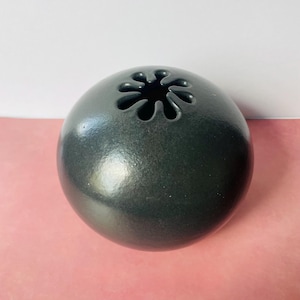 May include: A dark gray, spherical ceramic vase with a flower-shaped opening at the top. The vase has a smooth, glossy finish and sits on a pink surface. The design is simple and modern.