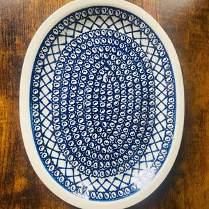 May include: An oval ceramic plate with a white rim and a blue and white geometric pattern. The pattern features concentric circles and diamond shapes. The plate is on a wooden surface.