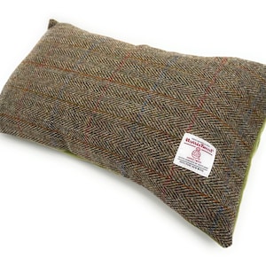 May include: A rectangular pillow with a brown and green herringbone tweed pattern. The pillow has a label that reads "Harris Tweed" and "100% Pure New Wool". The pillow is a neutral color and would fit in any room.