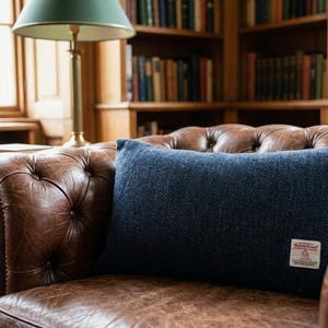May include: A navy blue rectangular pillow with a herringbone pattern rests on a brown leather armchair. The pillow has a small rectangular label on the front. A wooden bookshelf and a table lamp are in the background.
