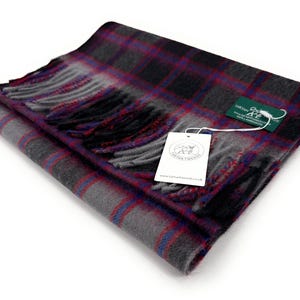 May include: A folded grey, black, and purple tartan scarf with fringed edges. The scarf has a tag attached with white string. The tartan pattern includes red and blue stripes. The scarf is made of a soft material.