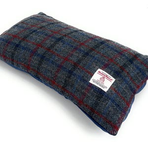 May include: Rectangular pillow with a gray, blue, and red plaid pattern. The pillow has a blue underside and a white label with red text. The label reads "Harris Tweed" and "100% Pure New Wool".