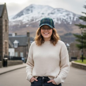 May include: A woman wearing a cream-colored sweater, dark jeans, and a plaid baseball cap. The cap has a green, navy, and yellow pattern. The background includes a snow-covered mountain and a street lined with buildings.
