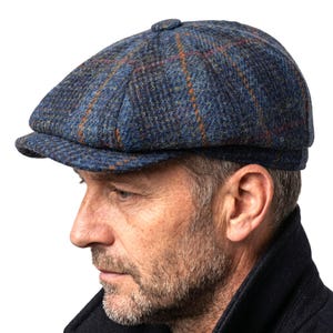 May include: A blue tweed newsboy cap with a plaid pattern in orange and red. The cap has a small brim and a button on top. The hat is made of a textured fabric.