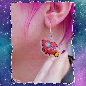 Könnte beinhalten: Ein Paar transparente Ohrringe aus Kunststoff mit einem roten Raketenmotiv. Die Rakete hat ein weißes Fenster und den Text "Alpiste" darauf gedruckt.